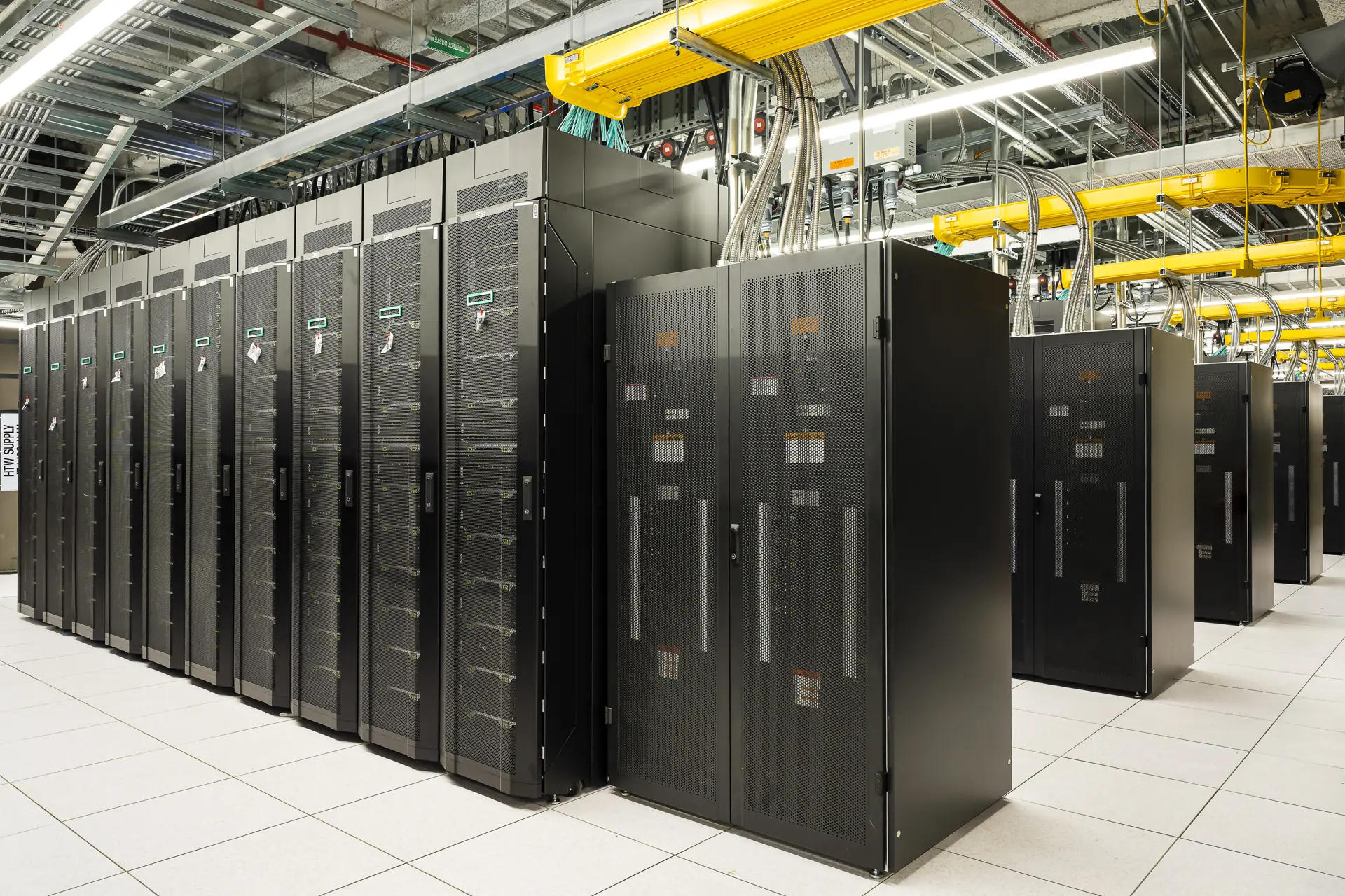 Rows of liquid-cooled supercomputer cabinets with overhead coolant and power distribution in a large data hall